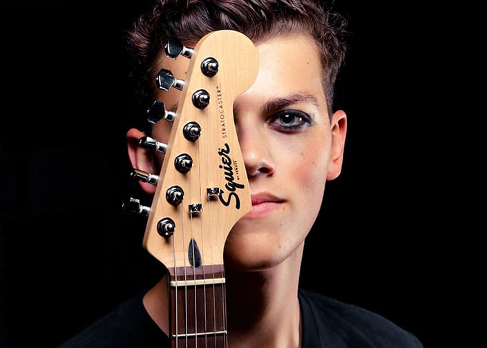 Person with guitar over shoulder against dark background.