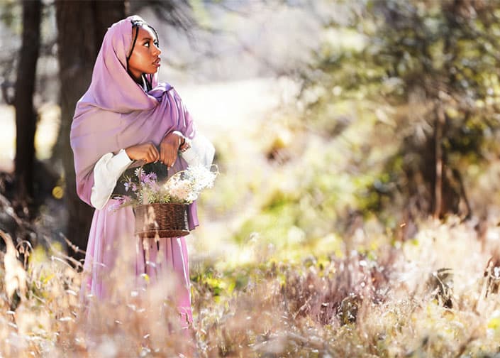 Person in purple hijab holding a basket of flowers in a sunny forest meadow.