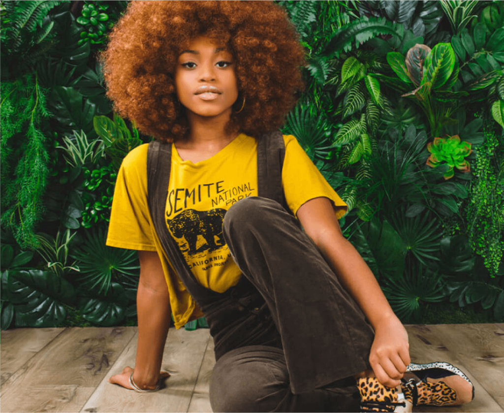 Person in yellow shirt and brown pants kneeling in front of a lush green plant backdrop.