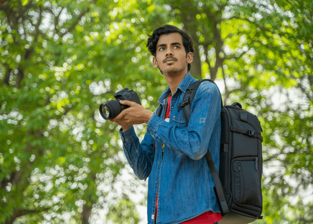 A person with a camera and backpack standing amidst green foliage.
