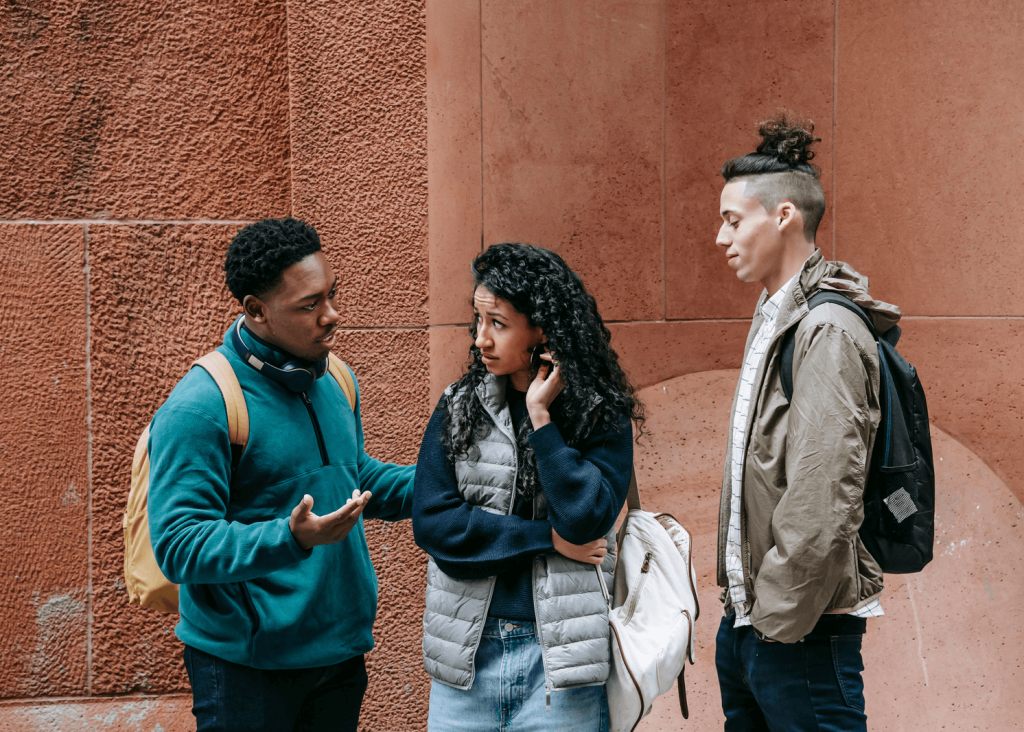 Three young individuals with backpacks stand in front of a textured wall.