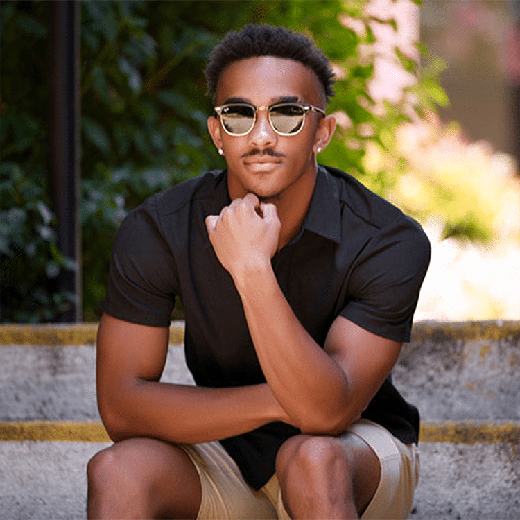 Man in a black shirt sitting on outdoor steps with lush greenery in the background.
