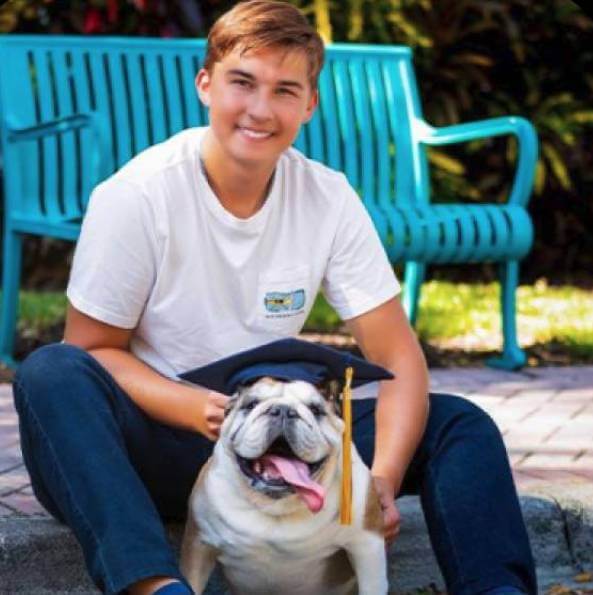 Person sitting with a bulldog wearing a graduation cap, holding a diploma, near a blue bench.